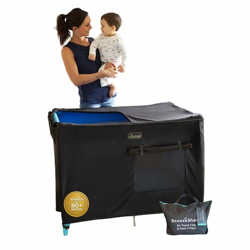 SnoozeShade blackout cover fitted securely over a pack and play travel crib while a mother holds her baby next to it.